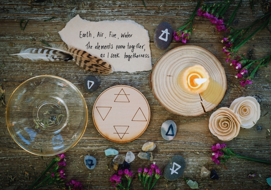 This image features a wooden tabletop arranged with various spiritual and natural elements, including:  A handwritten note that reads: "Earth, Air, Fire, Water. The elements come together, as I seek togetherness." A wooden disc engraved with symbols representing the four classical elements. A burning candle placed on another wooden slice, emitting a soft glow. Small stones and crystals, some etched with elemental symbols. Feathers, sprigs of purple flowers, and other organic materials. A clear glass bowl, possibly for use in rituals or as a vessel for water. The overall arrangement has a harmonious, ritualistic feel, blending natural and symbolic elements to evoke connection and balance.
